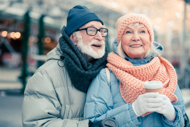 Two cute pensioners in warm winter clothes standing in the street with cups of coffee and smiling happily while looking away